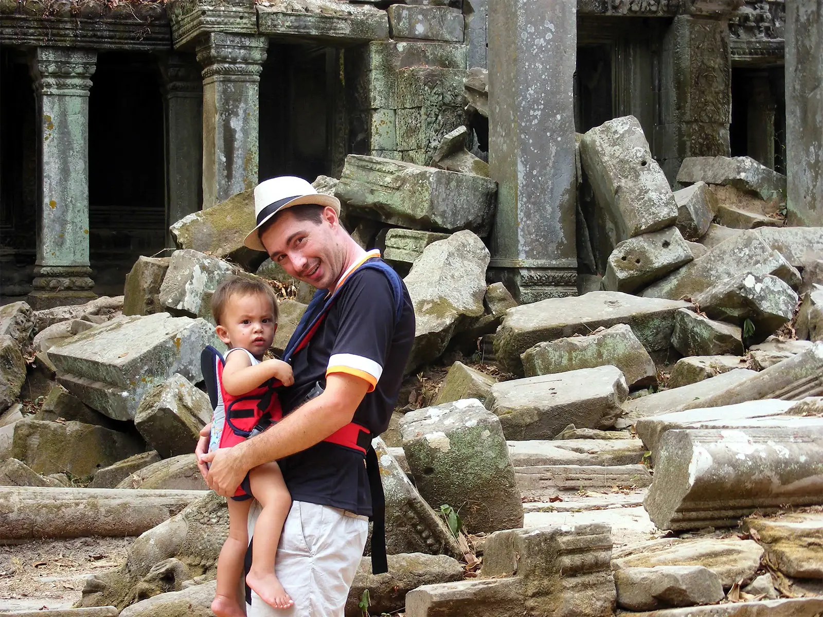Un voyageur portant un bébé en porte-bébé parmi les blocs écroulés d’un temple d’Angkor