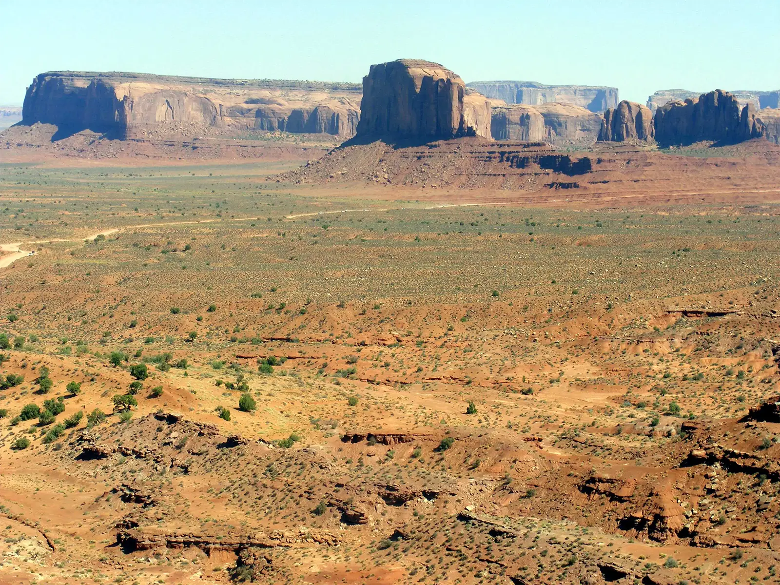 Plaine désertique parsemée d’arbustes et buttes de grès à l’horizon de Monument Valley