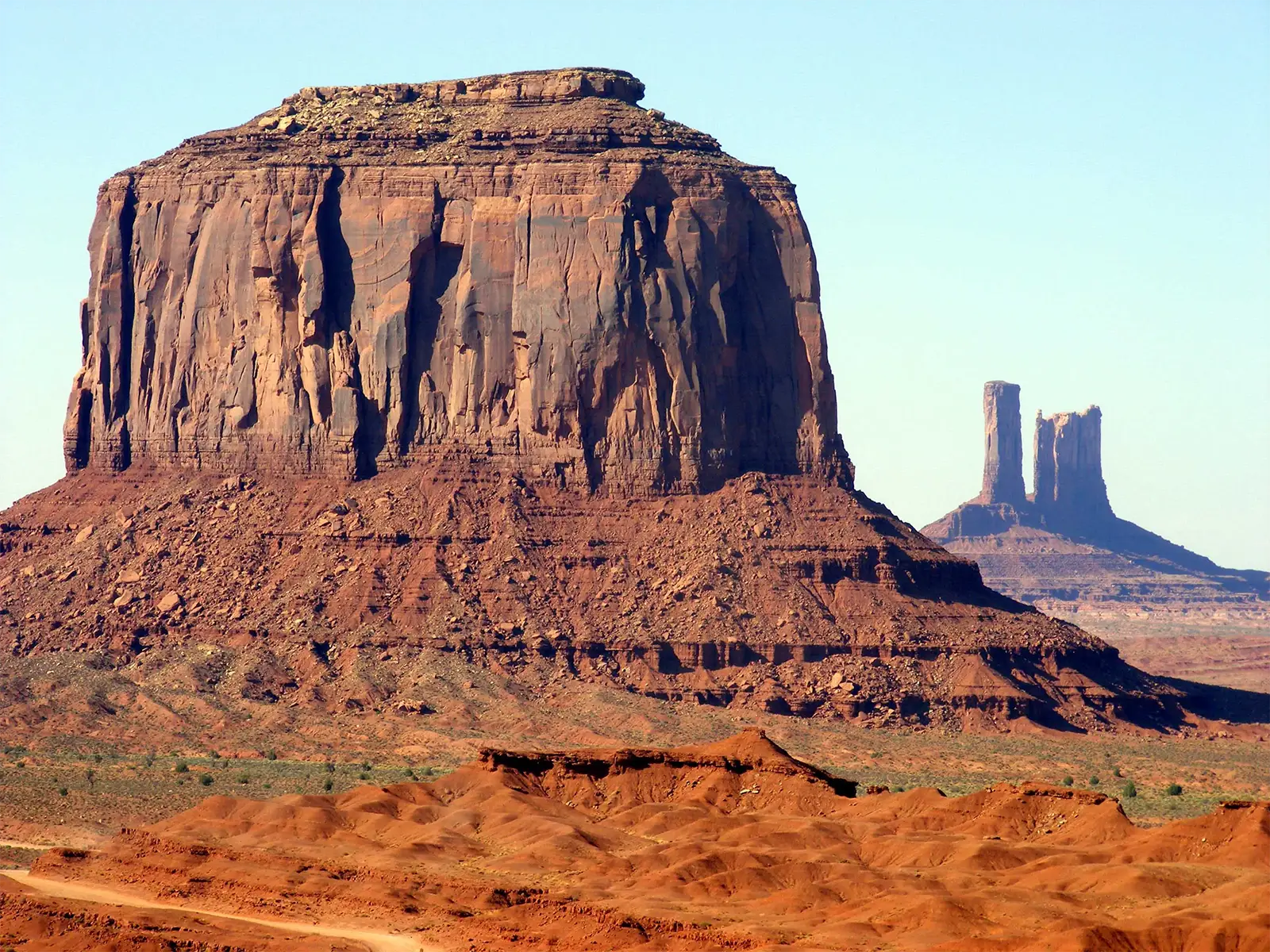 Imposante butte de grès rouge et pinacles jumeaux au loin dans la lumière du matin à Monument Valley