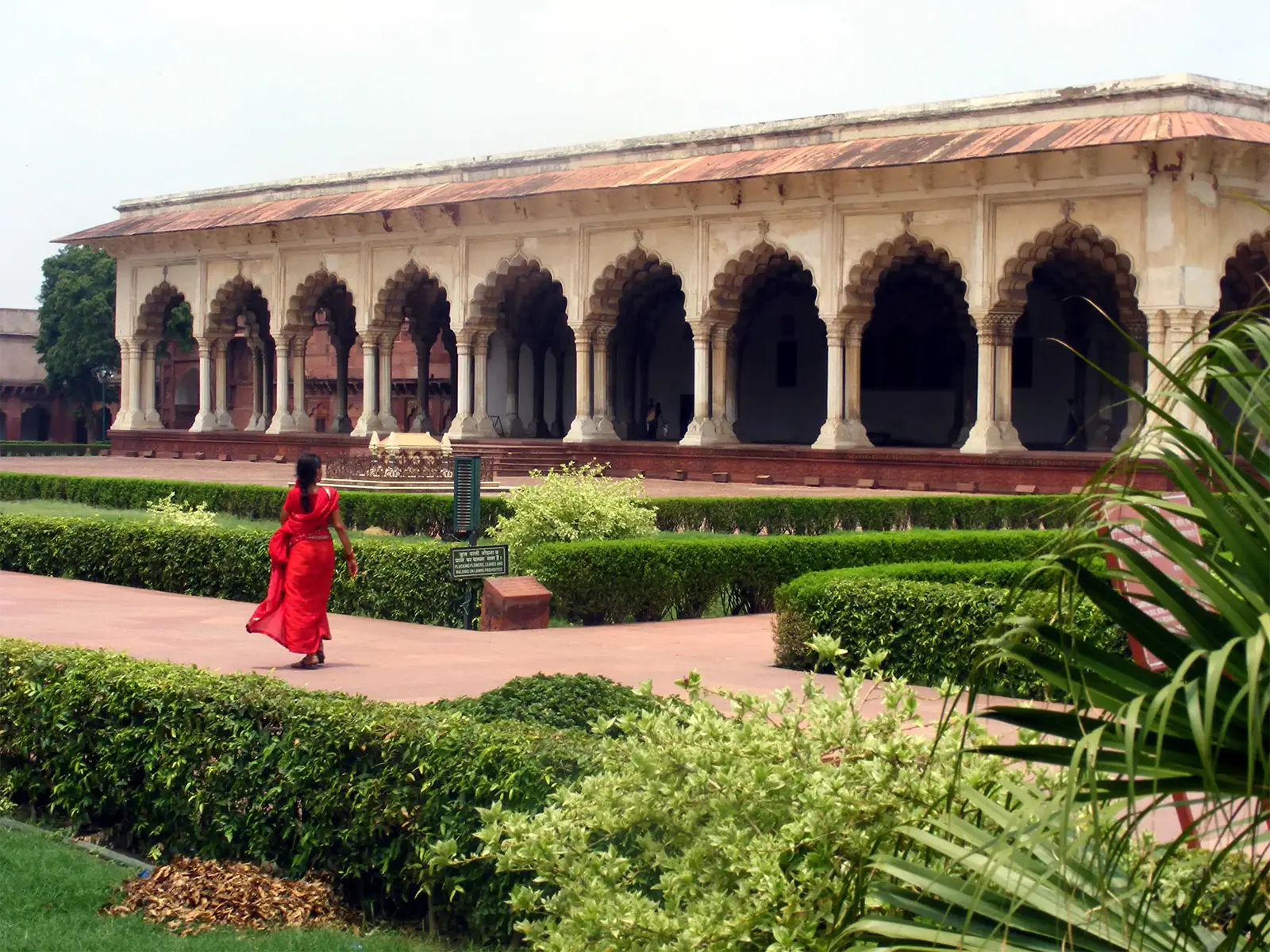 Femme en sari rouge dans les jardins, devant une salle à arcades du fort d’Agra