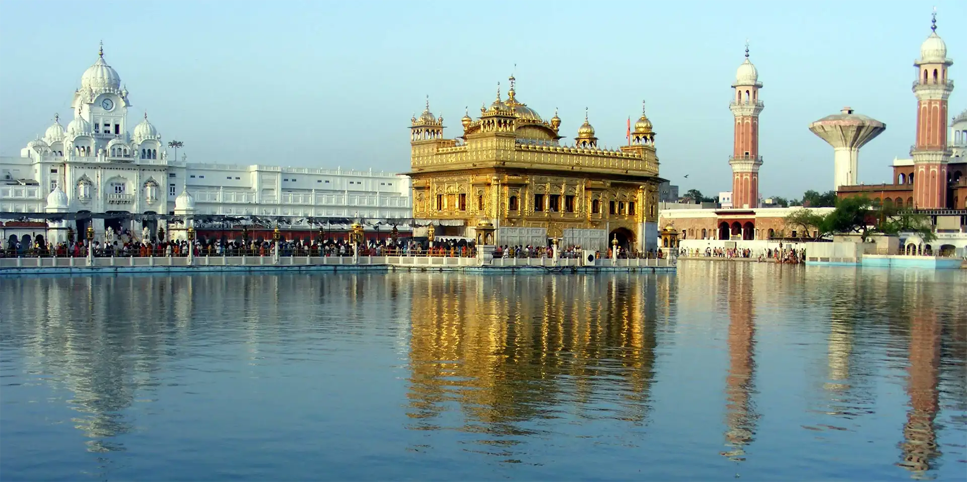 Panorama du Temple d’Or d’Amritsar entre édifices blancs et tours rouges, reflété dans le bassin