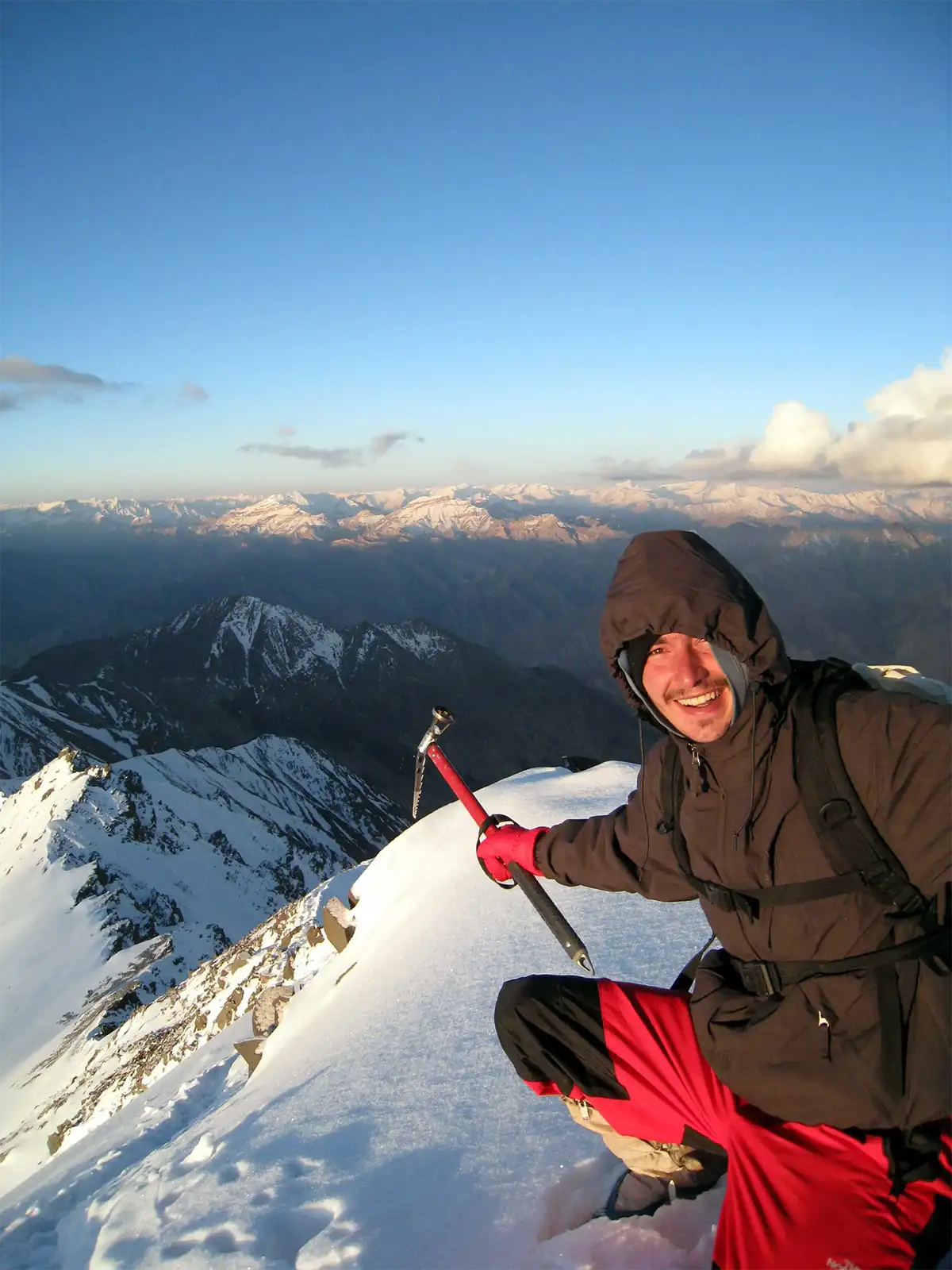 Un voyageur souriant accroupi avec son piolet au sommet enneigé du Stok Kangri