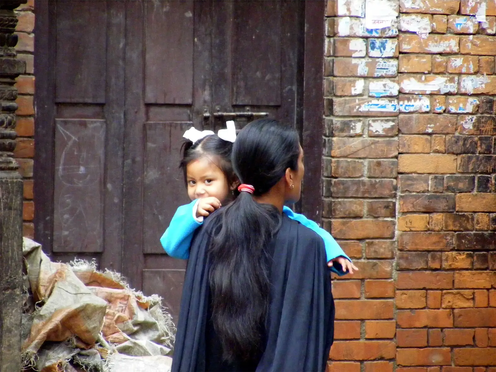 Mère portant sa fillette aux nœuds blancs devant un mur de briques à Bhaktapur