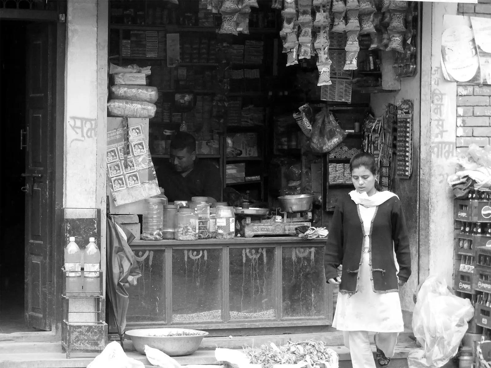 Épicier dans sa boutique et jeune femme passant devant, en noir et blanc à Bhaktapur