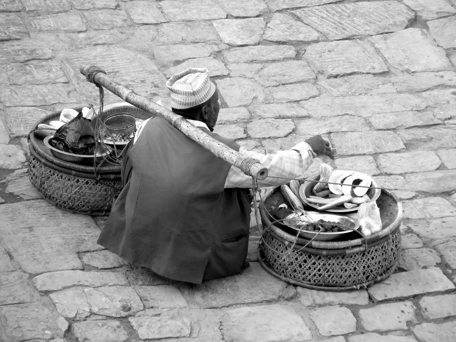 Marchand ambulant assis près de ses paniers en osier et de sa palanche, en noir et blanc à Bhaktapur