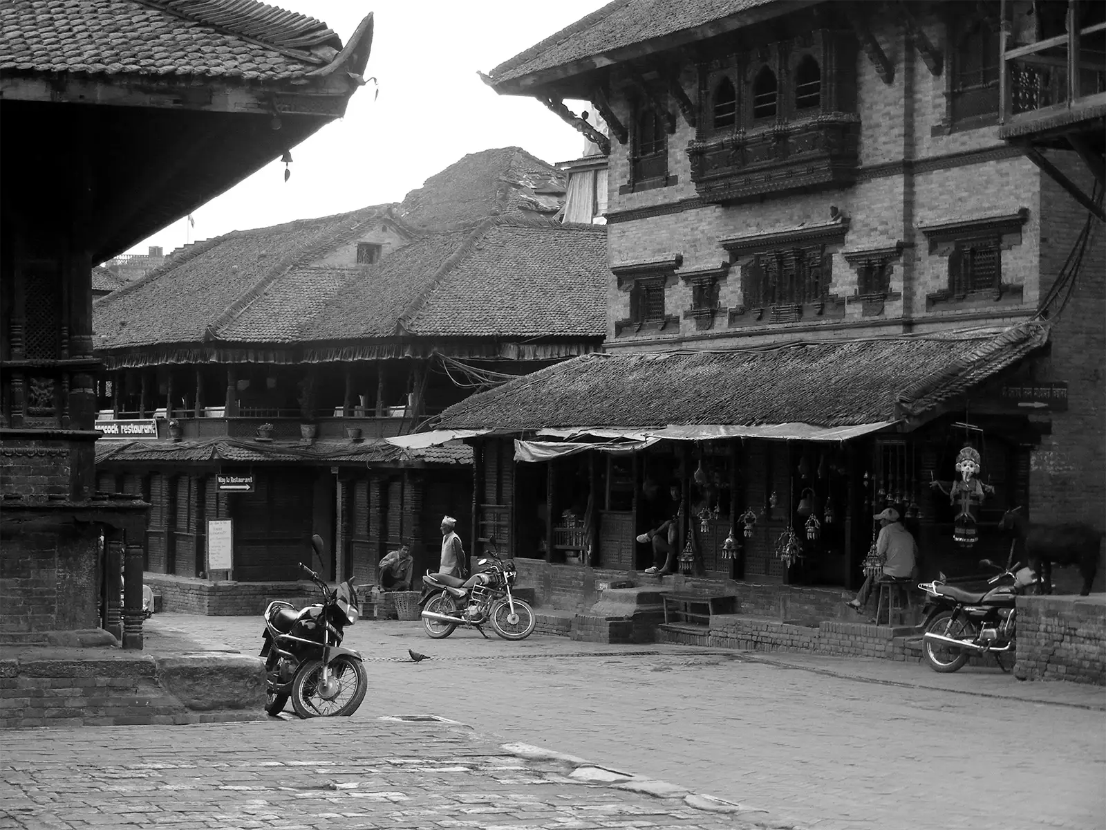 Place de Bhaktapur en noir et blanc, maisons anciennes, motos et échoppe de marionnettes