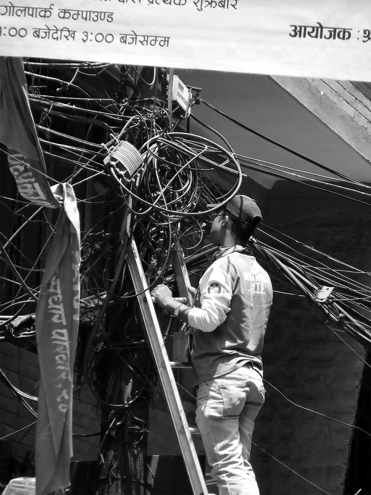 Électricien sur une échelle face à un enchevêtrement de câbles à Katmandou, en noir et blanc