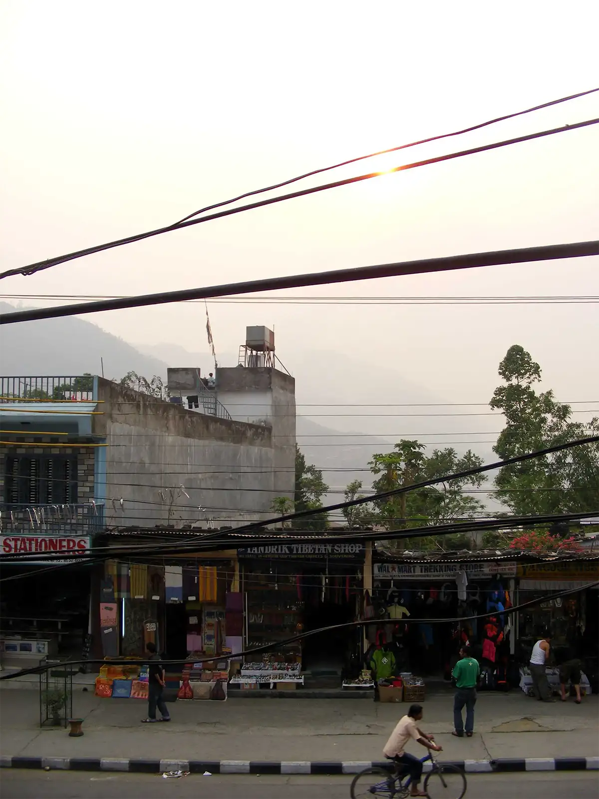 Boutiques tibétaines et cycliste dans une rue de Pokhara, soleil voilé derrière les câbles