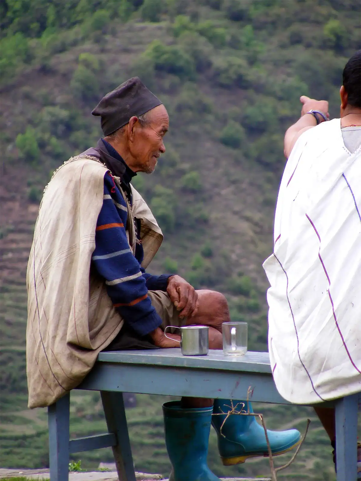 Vieil homme coiffé d’un topi buvant le thé sur un banc, face aux terrasses du trek des Annapurnas
