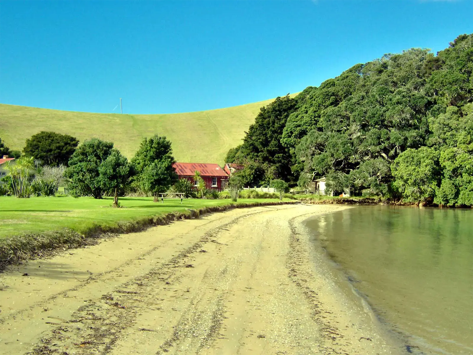 Plage de sable, maison au toit rouge et colline verte au bord d’une anse de la Bay of Islands