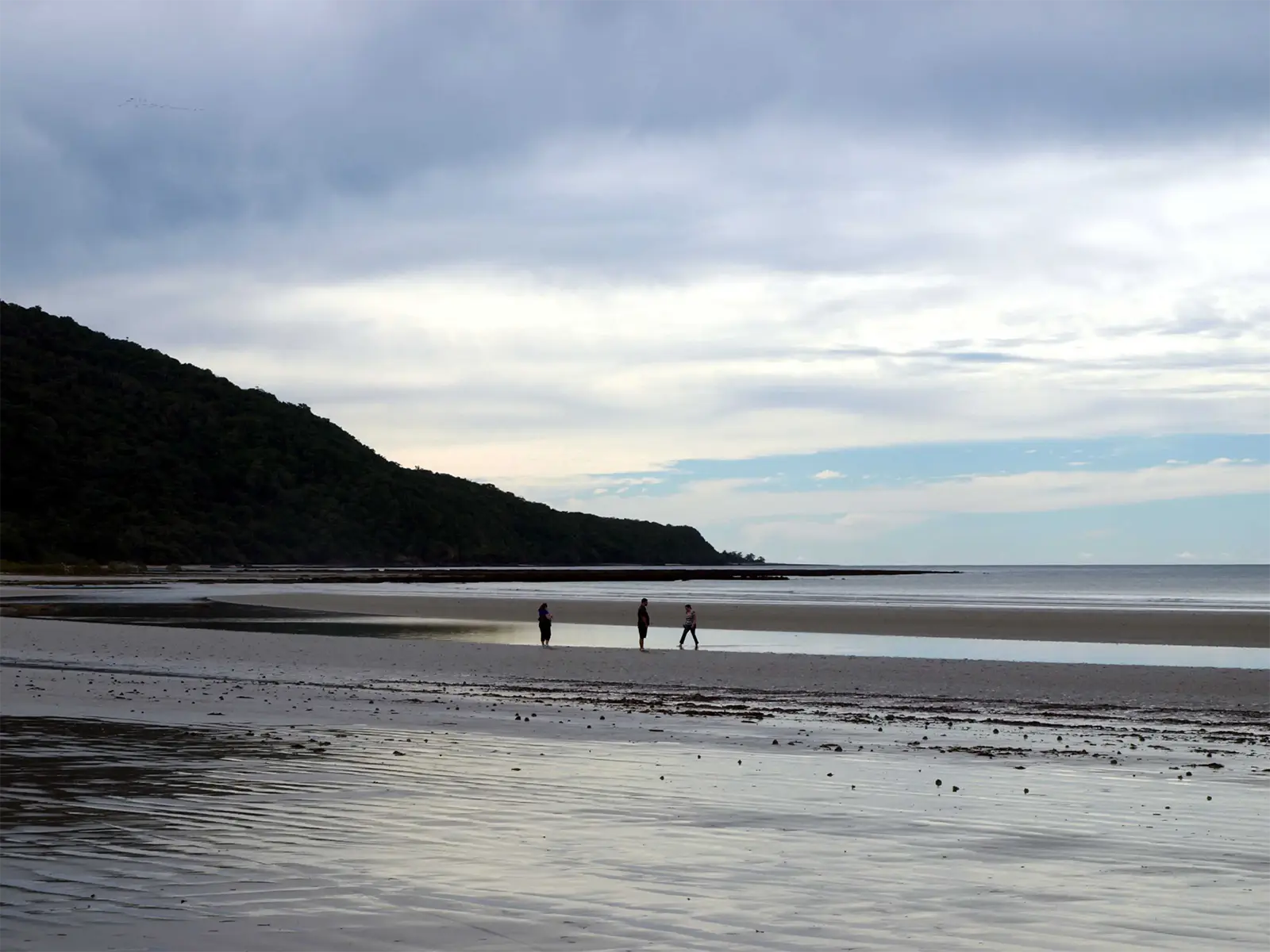 Promeneurs sur une large plage à marée basse sous un ciel nuageux, péninsule de Coromandel