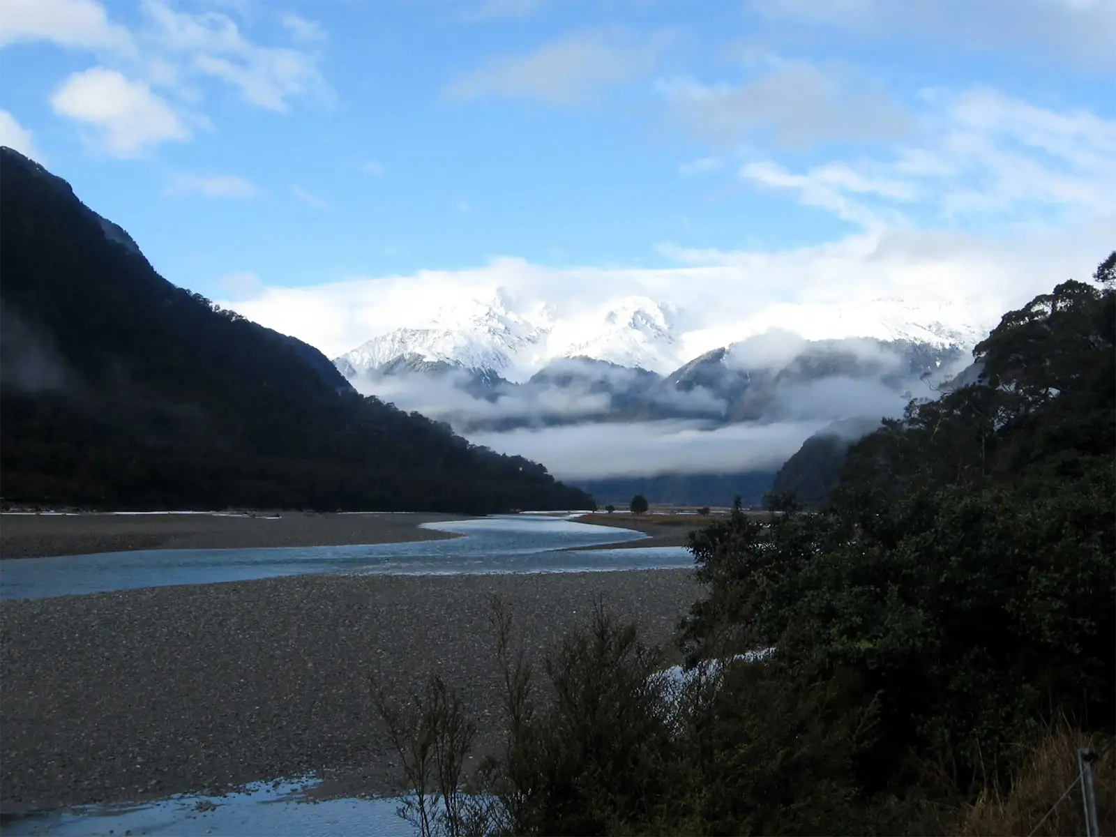 Rivière aux larges bancs de galets au pied de sommets enneigés dans les nuages, sur l’île du Sud