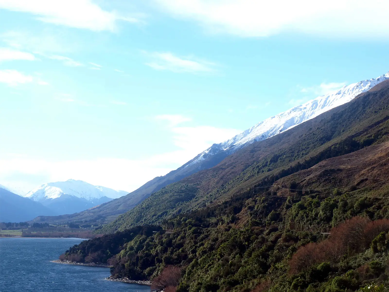 Lac bleu au pied de pentes boisées et de montagnes enneigées, sur la route de l’île du Sud