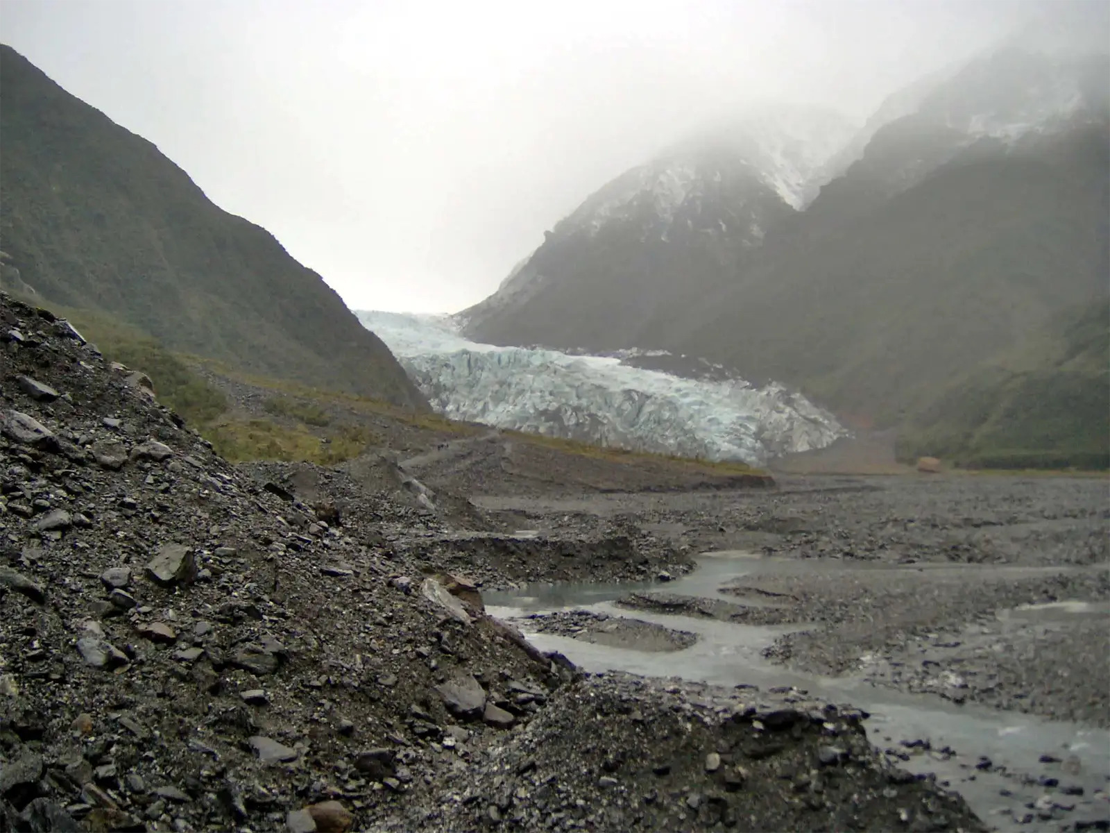 Front de glacier au fond d’une vallée de graviers et de torrents, sous la brume de la côte ouest