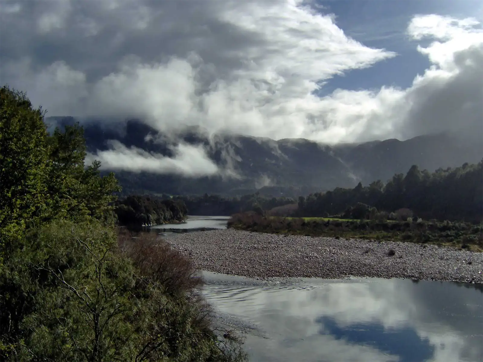 Rivière aux reflets du ciel et bancs de galets sous les nuages accrochés aux montagnes, vers la côte ouest