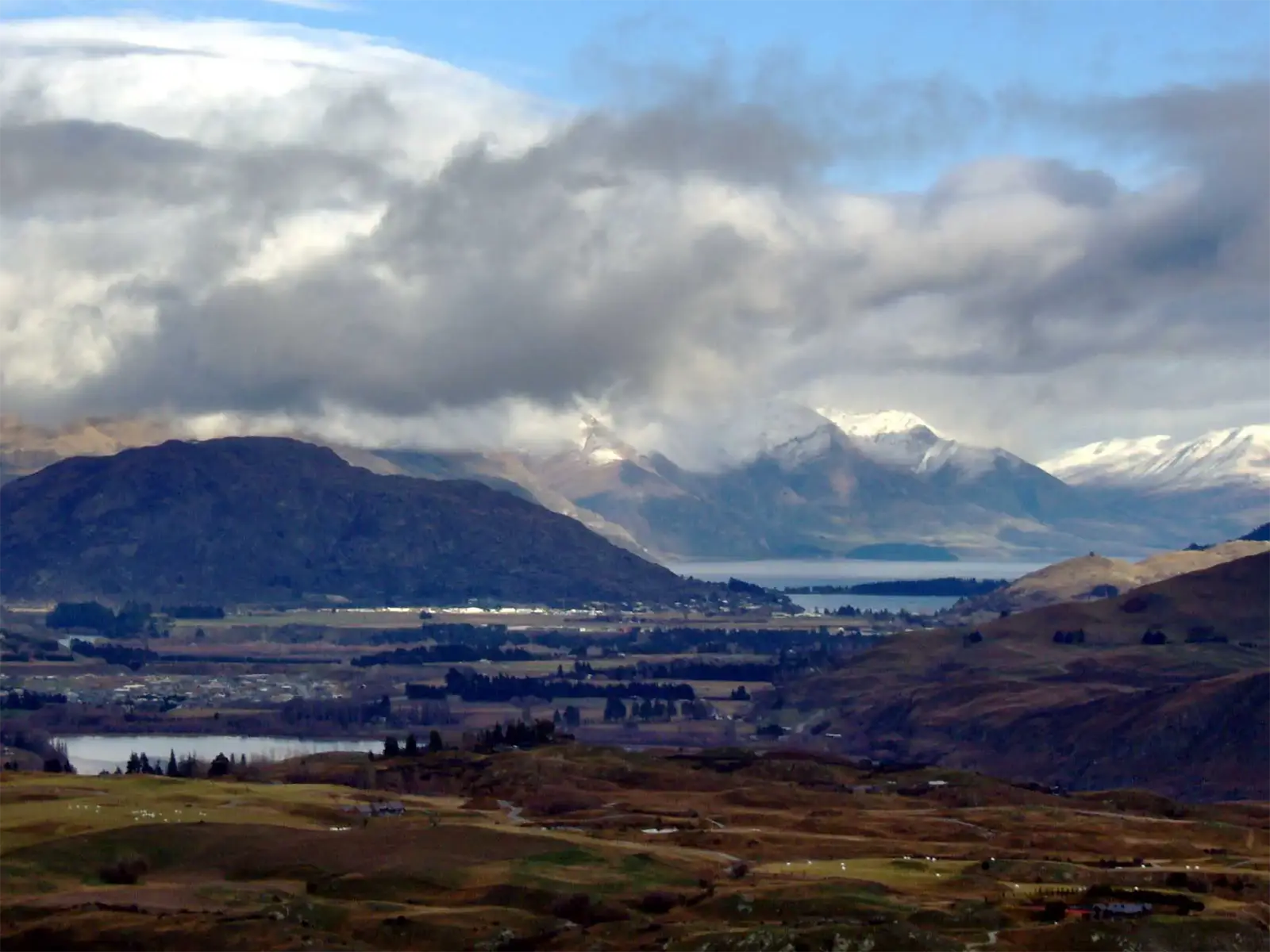 Lacs, plaine et sommets enneigés sous un ciel chargé, en panorama sur la région de Queenstown