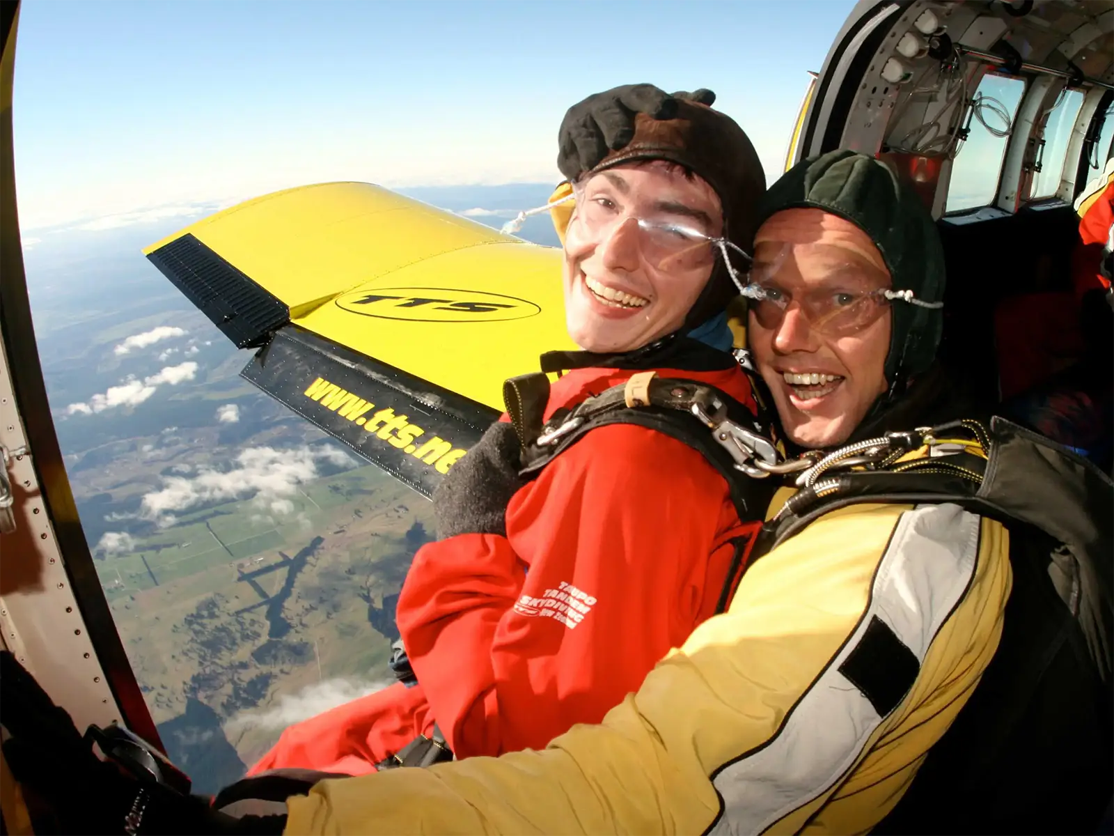 Deux parachutistes en tandem souriant à la porte de l’avion, au-dessus de la campagne de Taupo