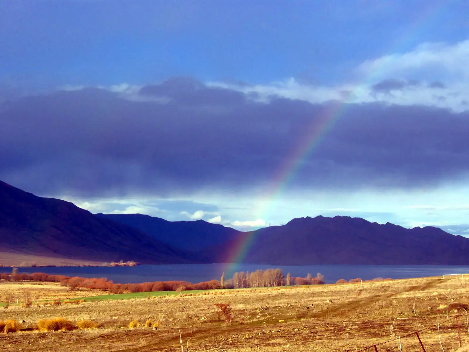 Arc-en-ciel au-dessus d’un lac et de montagnes sombres, sur la route du lac Tekapo