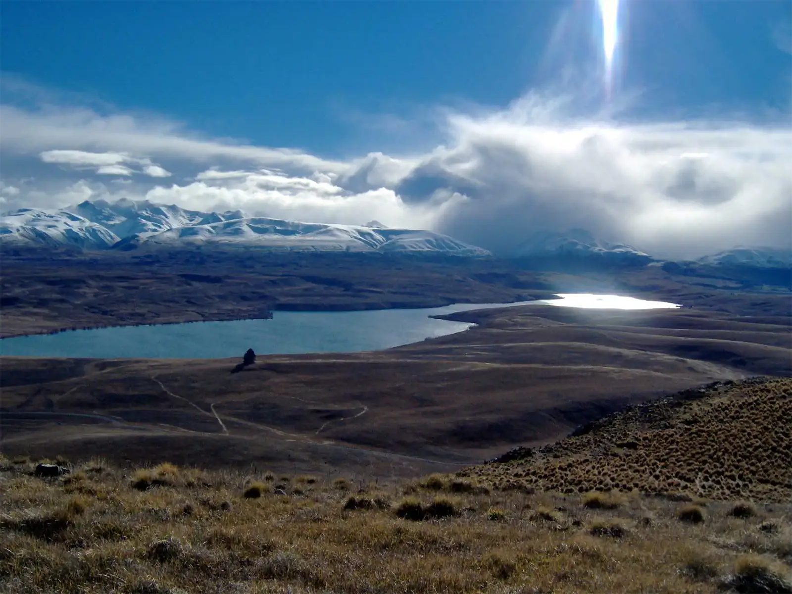 Le lac Alexandrina et les montagnes enneigées sous un rayon de soleil, vus des collines d’herbe sèche du mont John