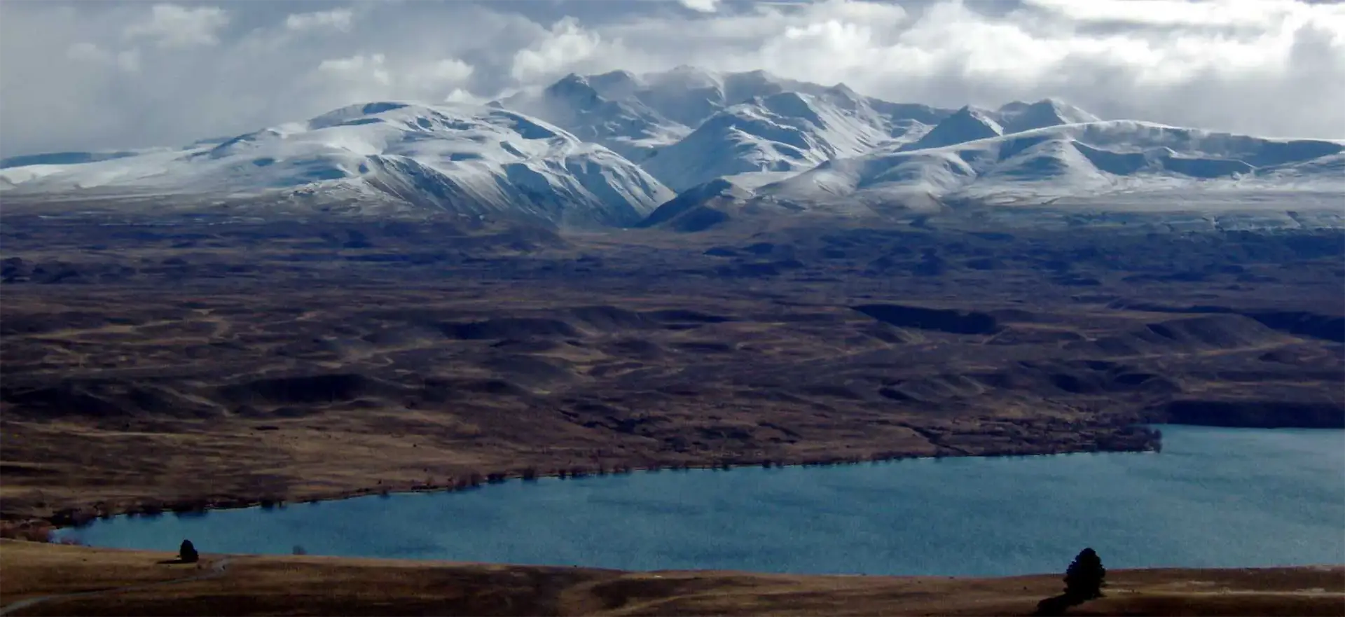 Panorama sur le lac Alexandrina et une plaine brune au pied des Alpes du Sud enneigées, vu du mont John à Tekapo