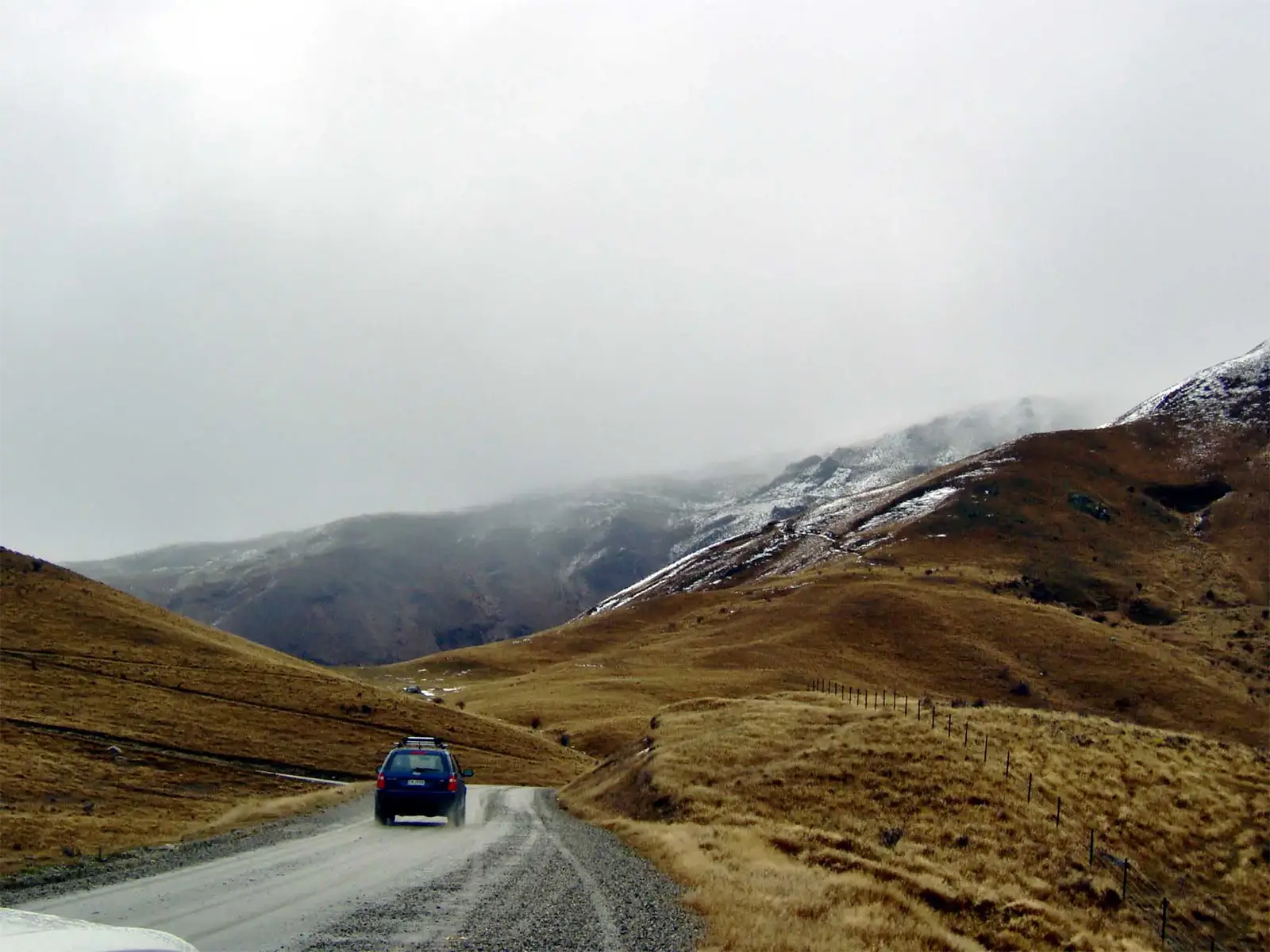 Route de gravier montant entre les collines d’herbe rase vers les sommets enneigés, près de Wanaka