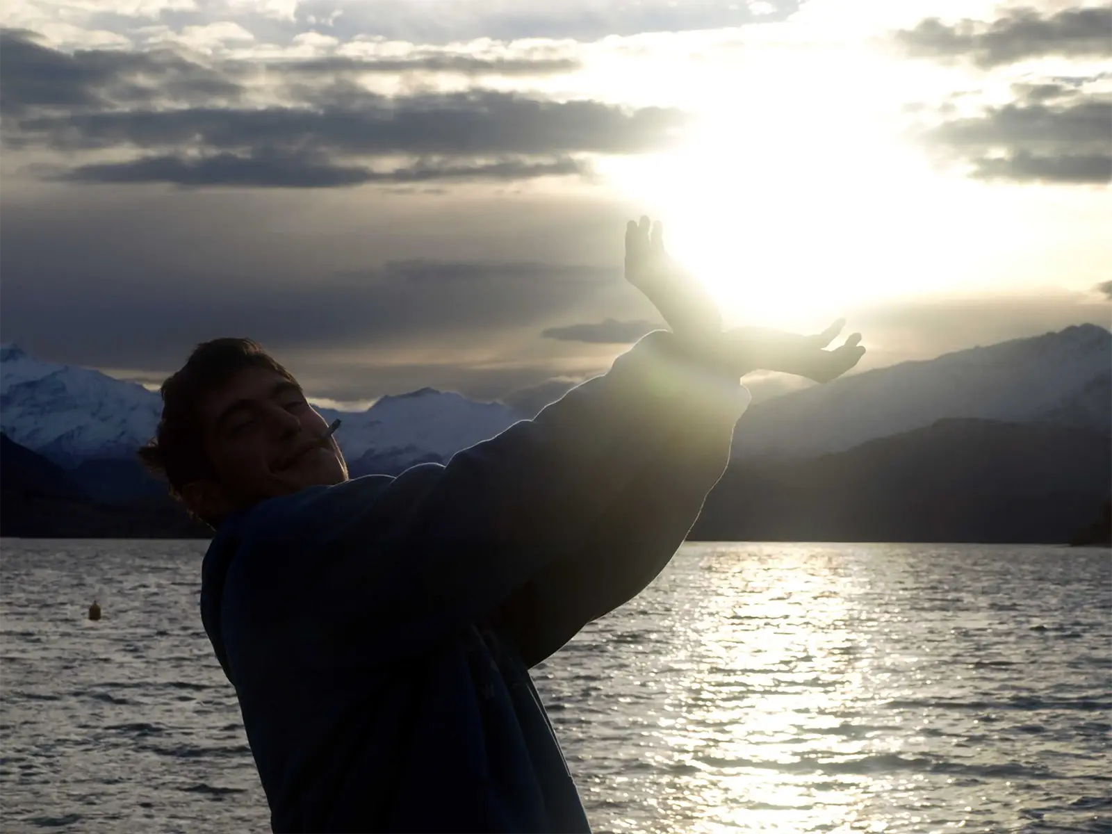 Un voyageur fait mine de tenir le soleil couchant au-dessus du lac Wanaka et des montagnes enneigées