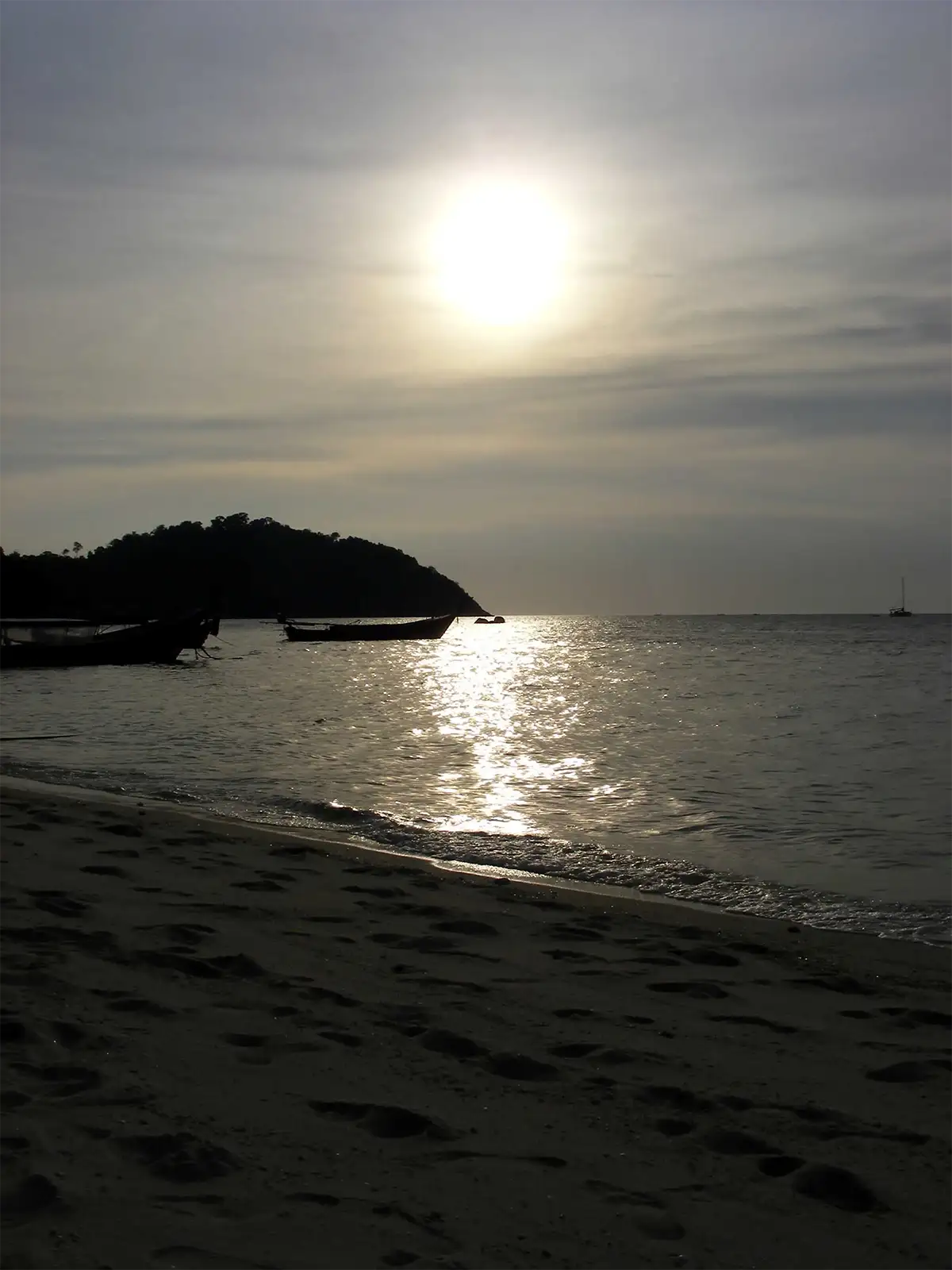 Soleil voilé se reflétant sur la mer devant une plage et des bateaux au mouillage à Koh Lipe