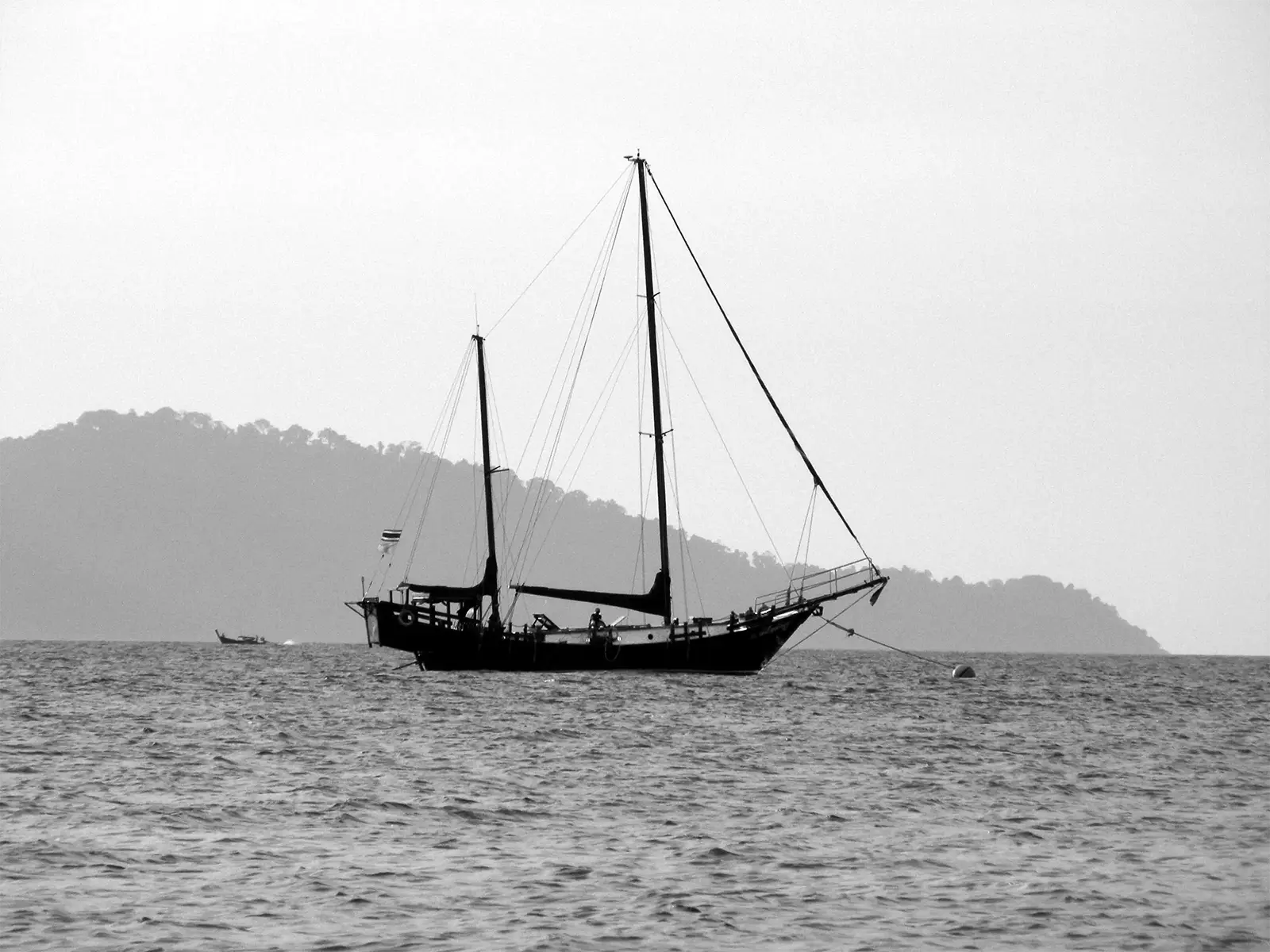 Voilier à deux mâts au mouillage devant une île boisée à Koh Lipe, en noir et blanc