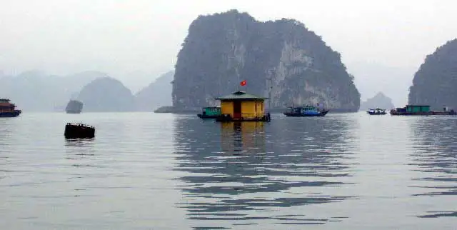 Maison flottante jaune et bateaux au pied d’un îlot karstique, dans la brume de la baie d’Halong